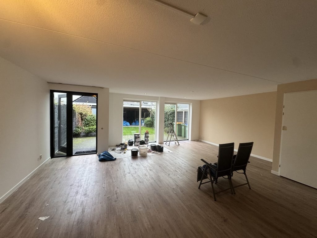 Woonkamer met lichte wanden, een houten vloer en klusspullen bij de tuindeuren